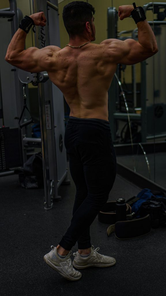 Adult male showcasing muscular physique in gym environment. Back view pose.